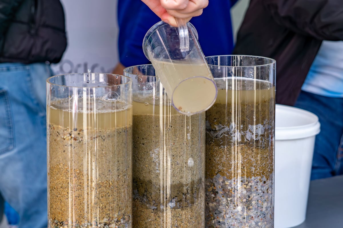 Water filtration demonstration using transparent columns filled with sand, gravel, and sediment. A person pours dirty water into one of the containers. An educational scene perfect for topics related to ecology, science, environmental protection